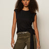 A woman with curly hair wears the Sanctuary Clothing side tie muscle tee in black, styled with olive green pants and a patterned belt, standing against a plain light background.