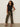 A woman with curly hair wears the Sanctuary Clothing side tie muscle tee in black, paired with olive green wide-leg pants, a patterned belt, and black heeled sandals, standing against a plain light background.
