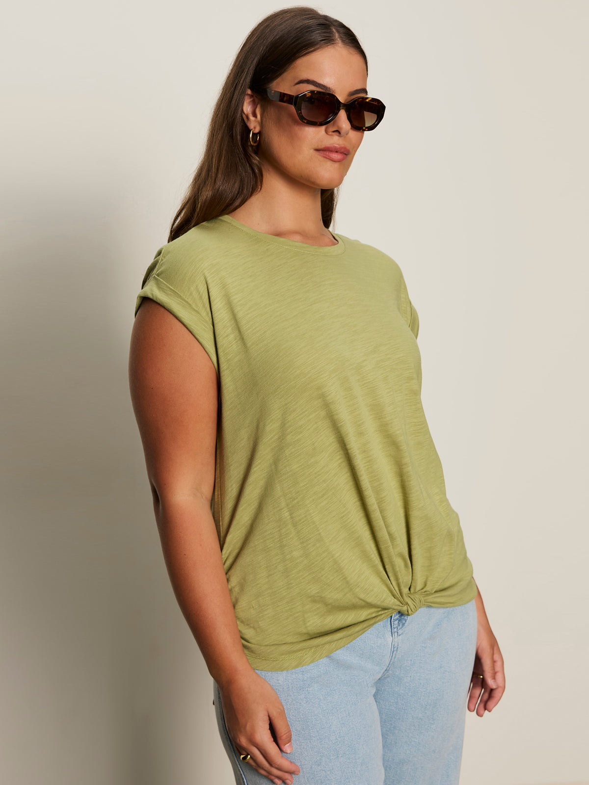 A woman in Sanctuary Clothing's knotted muscle tee bamboo extended sizing and light blue jeans stands against a plain background, facing slightly to the side with a neutral expression while wearing sunglasses.