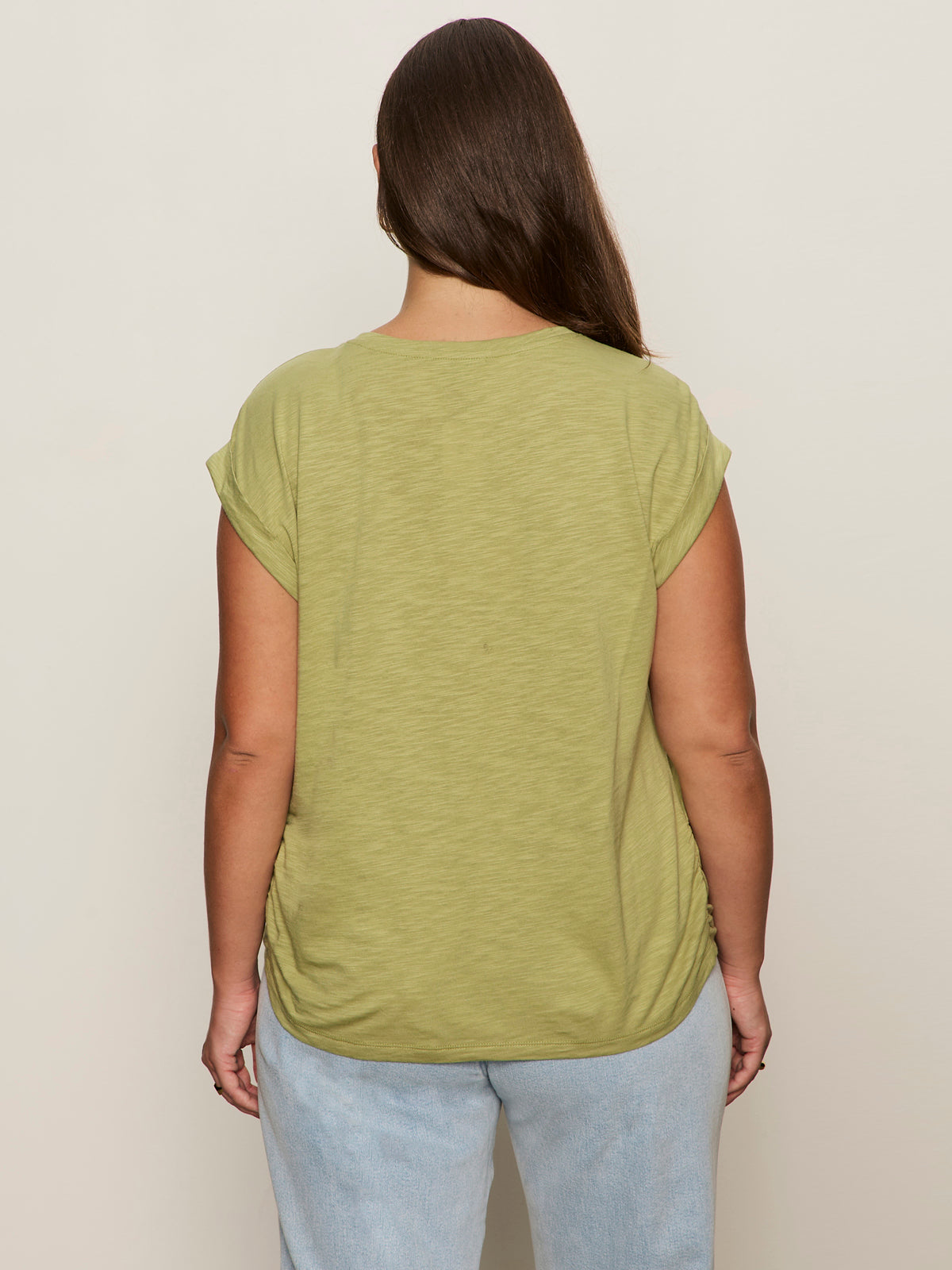 A woman with long brown hair stands facing away, wearing the Sanctuary Clothing knotted muscle tee bamboo extended sizing in light green and light blue jeans against a plain beige background.