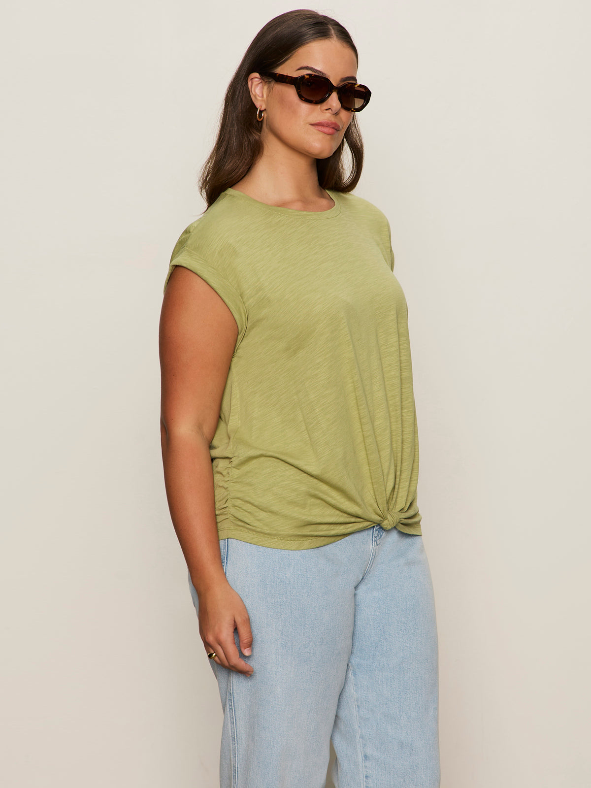 A woman wearing sunglasses, light blue jeans, and the Sanctuary Clothing knotted muscle tee bamboo (extended sizing) stands against a plain beige background.