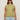 A woman wears dark sunglasses and the Sanctuary Clothing knotted muscle tee bamboo extended sizing in light olive green with light blue jeans, standing facing forward against a plain beige background.