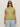 A woman wears dark sunglasses and the Sanctuary Clothing knotted muscle tee bamboo extended sizing in light olive green with light blue jeans, standing facing forward against a plain beige background.