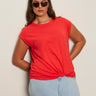 A woman wears the Sanctuary Clothing knotted muscle tee in cayenne (extended sizing) with light blue jeans. She stands against a light wall, has long brown hair, sunglasses, and her hands are behind her back.