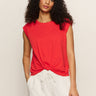 A woman with long curly hair wears the Sanctuary Clothing knotted muscle tee in cayenne and loose white pants, standing against a plain light background with her hands in her pockets.