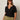 A woman with long brown hair wears sunglasses and the Sanctuary Clothing notch neck tee in black extended sizing, paired with light olive drawstring pants, standing against a plain light background.