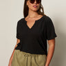 A woman with long brown hair wears sunglasses and the Sanctuary Clothing notch neck tee in black extended sizing, paired with light olive drawstring pants, standing against a plain light background.