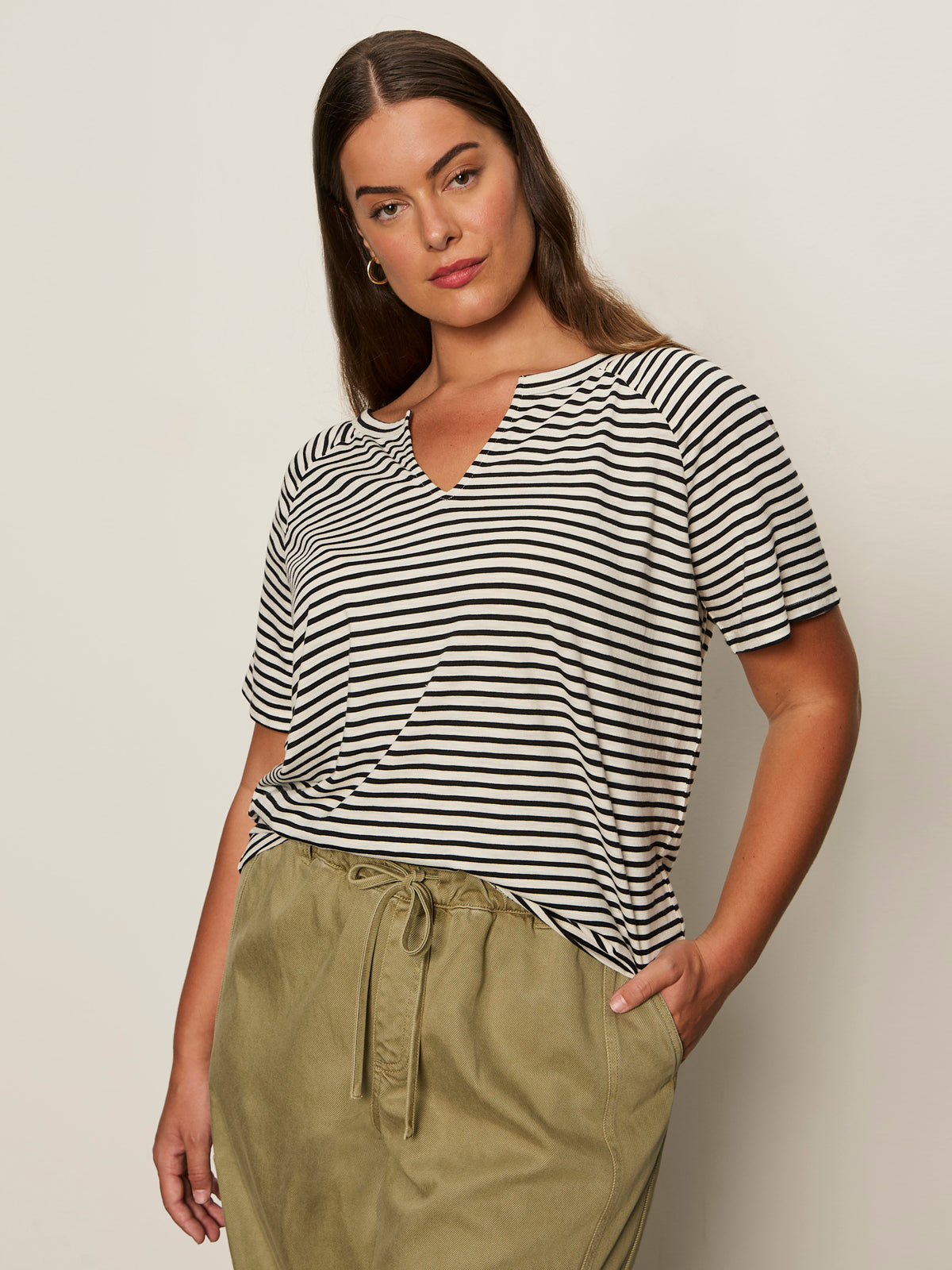 A woman with long brown hair models the Sanctuary Clothing notch neck tee light oat black stripe 3, paired with olive green drawstring pants, standing with one hand in her pocket against a plain light background.
