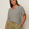 A woman with long brown hair models the Sanctuary Clothing notch neck tee light oat black stripe 3, paired with olive green drawstring pants, standing with one hand in her pocket against a plain light background.