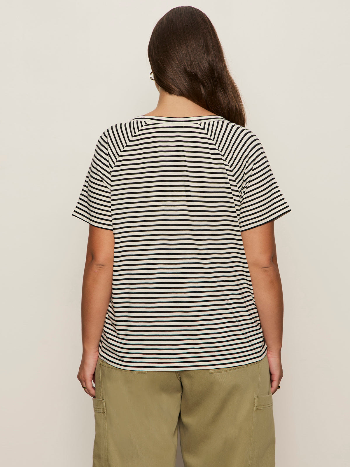 A person with long brown hair wears Sanctuary Clothing's notch neck tee in light oat black stripe, paired with olive green pants, standing against a plain light background.