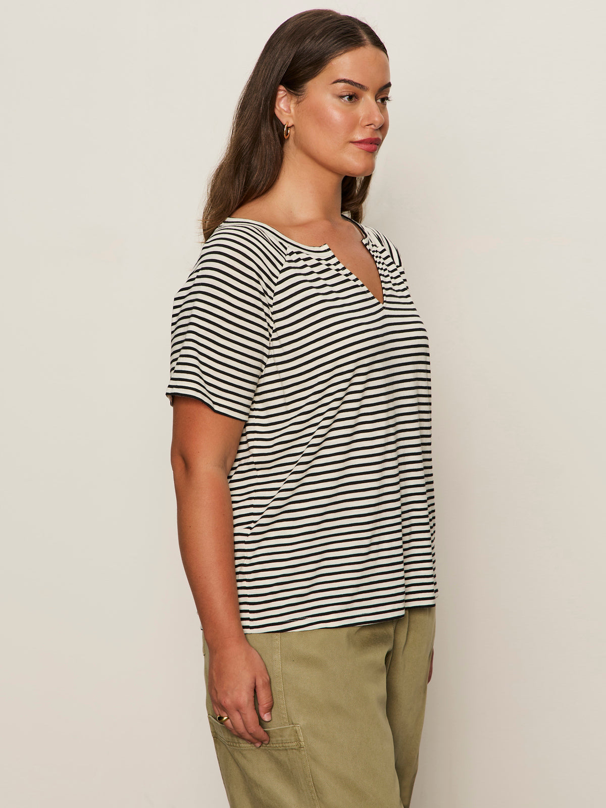 A woman with long brown hair and a neutral expression stands in profile against a plain background, wearing Sanctuary Clothing's notch neck tee light oat black stripe 3 and olive green pants.