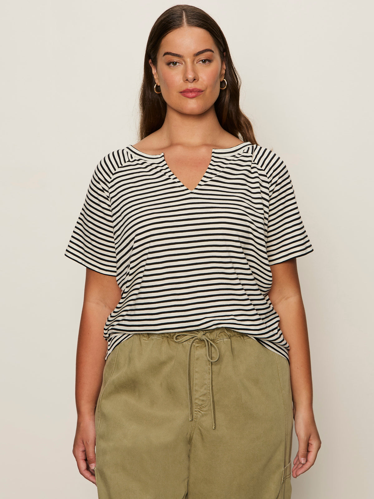 A woman with long brown hair wears the Sanctuary Clothing notch neck tee in light oat black stripe, paired with olive green drawstring pants, standing against a plain light background.