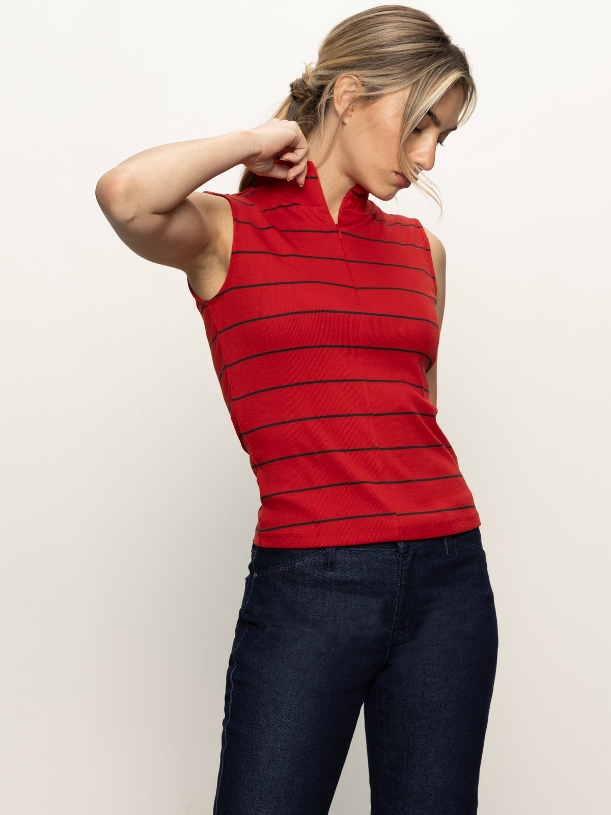 A woman wearing the Sanctuary Clothing sleeveless zip mock top in red energy with black stripes and dark blue jeans stands against a plain light background, looking down with one hand touching her neck.