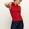A woman wearing the Sanctuary Clothing sleeveless zip mock top in red energy with black stripes and dark blue jeans stands against a plain light background, looking down with one hand touching her neck.