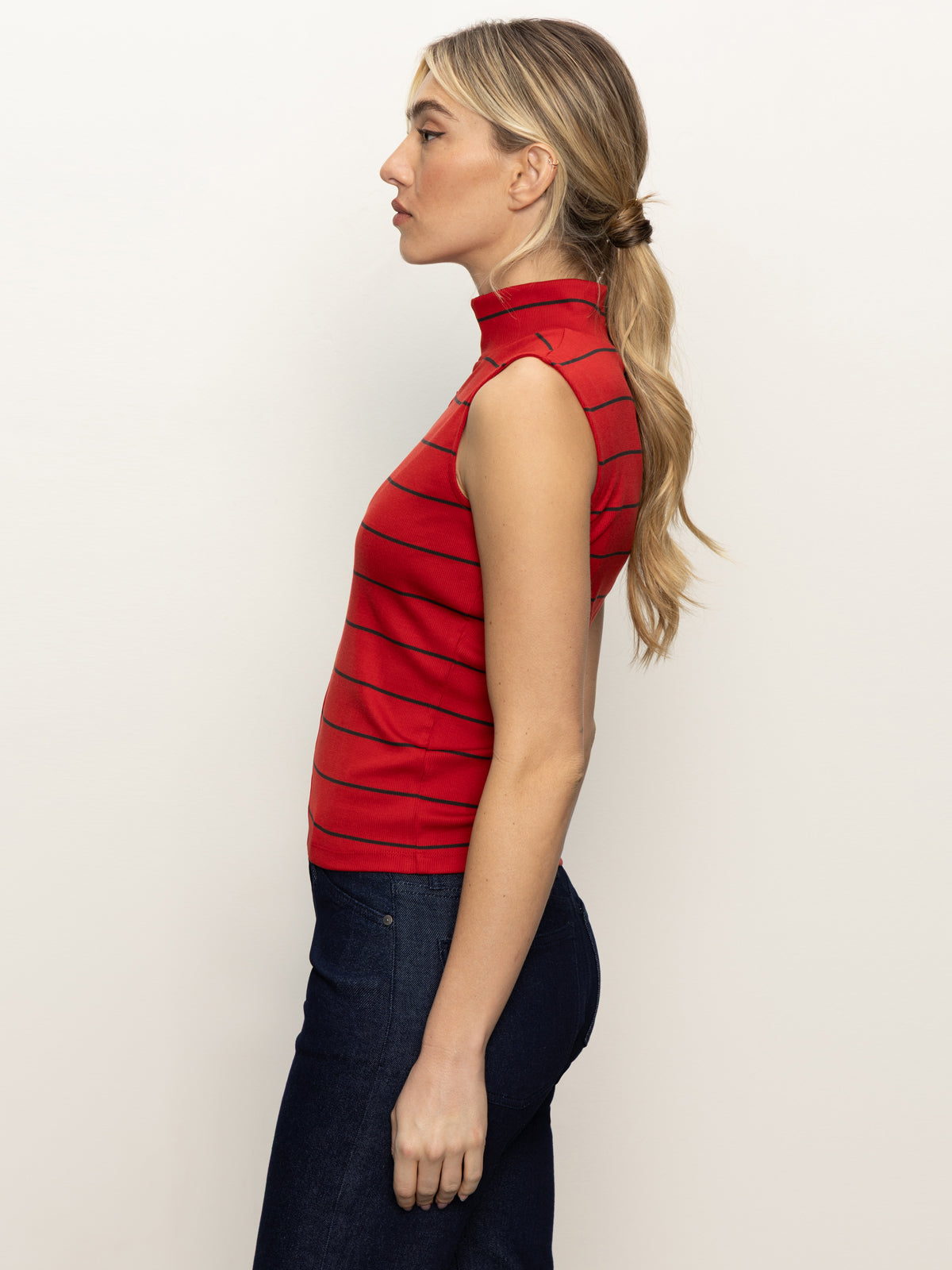 A woman in profile wears the Sanctuary Clothing sleeveless zip mock top in red energy black stripe and dark blue jeans. Her long blonde hair is tied in a low ponytail, and she stands against a plain background.