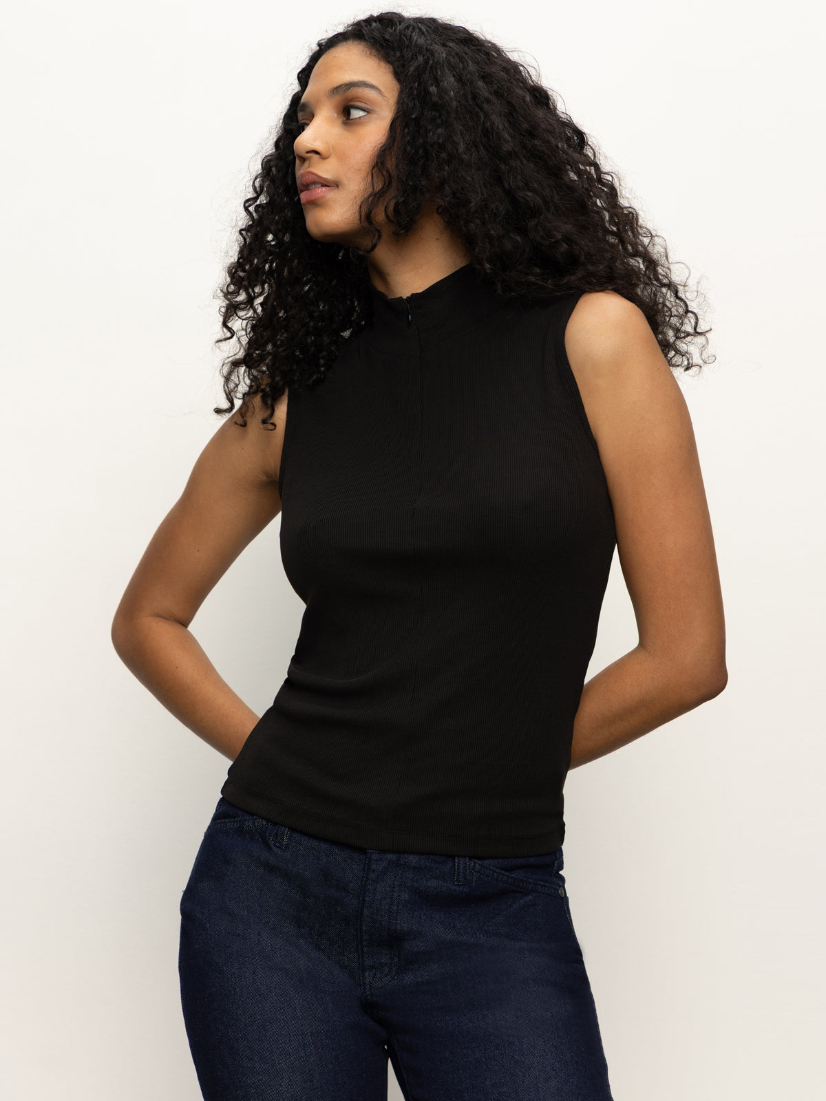 A woman with long curly hair wears the Sanctuary Clothing sleeveless zip mock top in black and dark jeans, standing against a plain light background, looking to her left with her hands at her sides.
