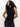 A woman with long curly hair wears the Sanctuary Clothing sleeveless zip mock top in black and dark jeans, standing against a plain light background, looking to her left with her hands at her sides.