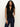 A woman with long curly hair models the Sanctuary Clothing sleeveless zip mock top in black, paired with dark blue jeans. She stands against a plain light background with one hand in her pocket.