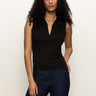 A woman with long curly hair models the Sanctuary Clothing sleeveless zip mock top in black, paired with dark blue jeans. She stands against a plain light background with one hand in her pocket.
