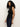 A woman with long curly hair wears the Sanctuary Clothing sleeveless zip mock top in black, paired with dark blue jeans, and stands against a plain light background looking at the camera with a neutral expression.