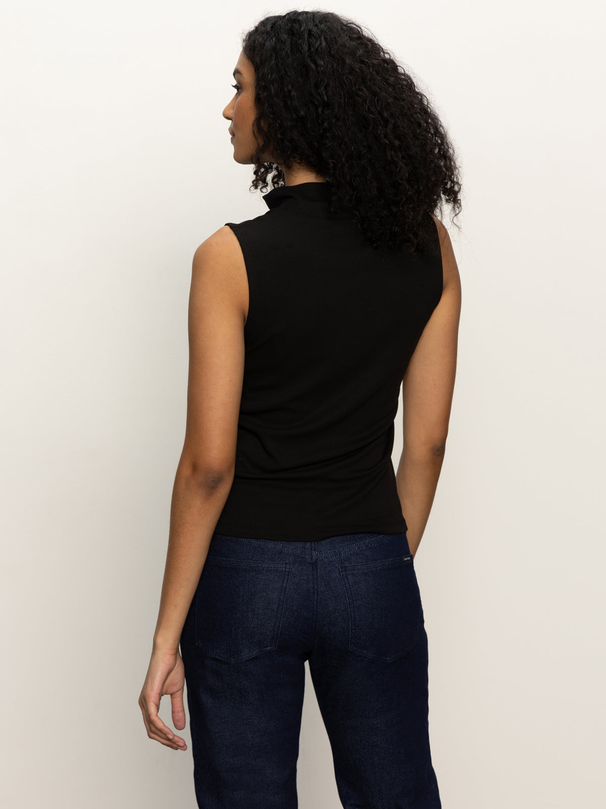 A person with curly hair faces away from the camera, wearing the Sanctuary Clothing sleeveless zip mock top in black and dark blue jeans, set against a plain, light-colored background.