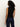 A person with curly hair faces away from the camera, wearing the Sanctuary Clothing sleeveless zip mock top in black and dark blue jeans, set against a plain, light-colored background.