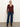 A woman with blonde hair tied back and a neutral expression stands against a white wall, wearing the Sanctuary Clothing long sleeve textured top in chicory, blue cuffed wide-leg jeans, and black heeled boots.