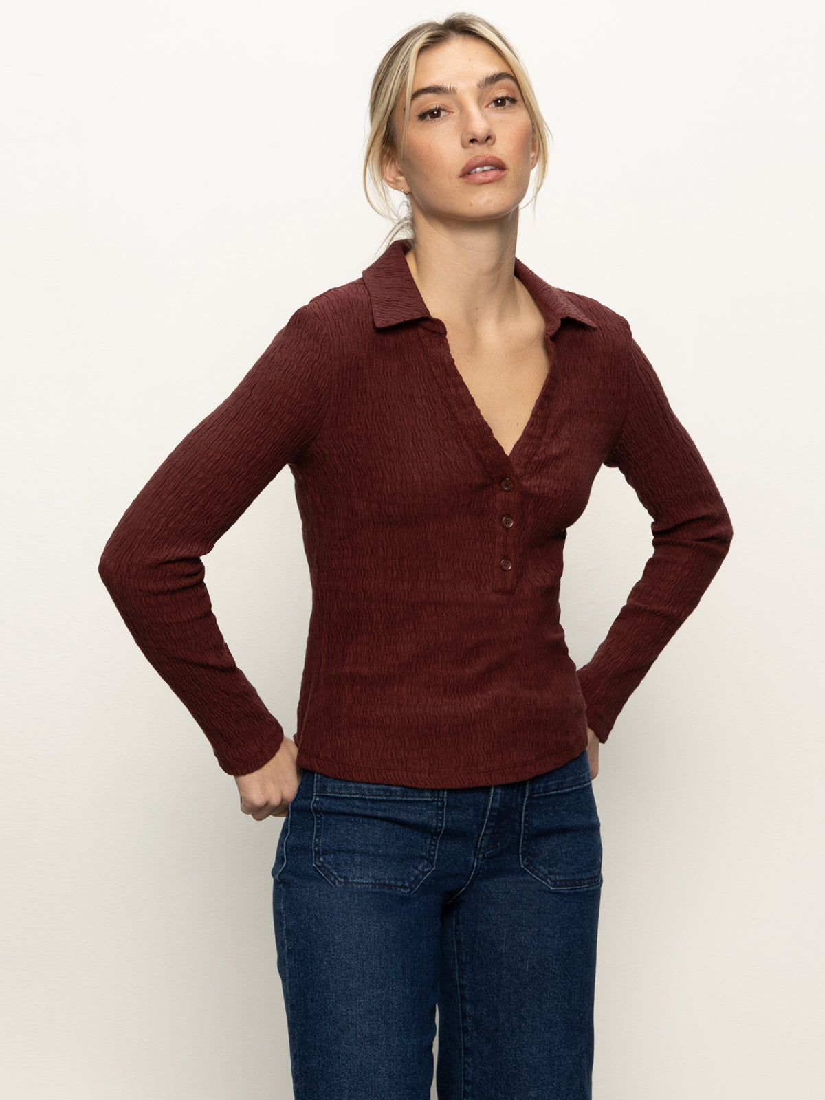 A woman wears the Sanctuary Clothing long sleeve textured top in chicory with jeans, standing against a plain light background, gazing slightly upward and resting her hands on her hips.