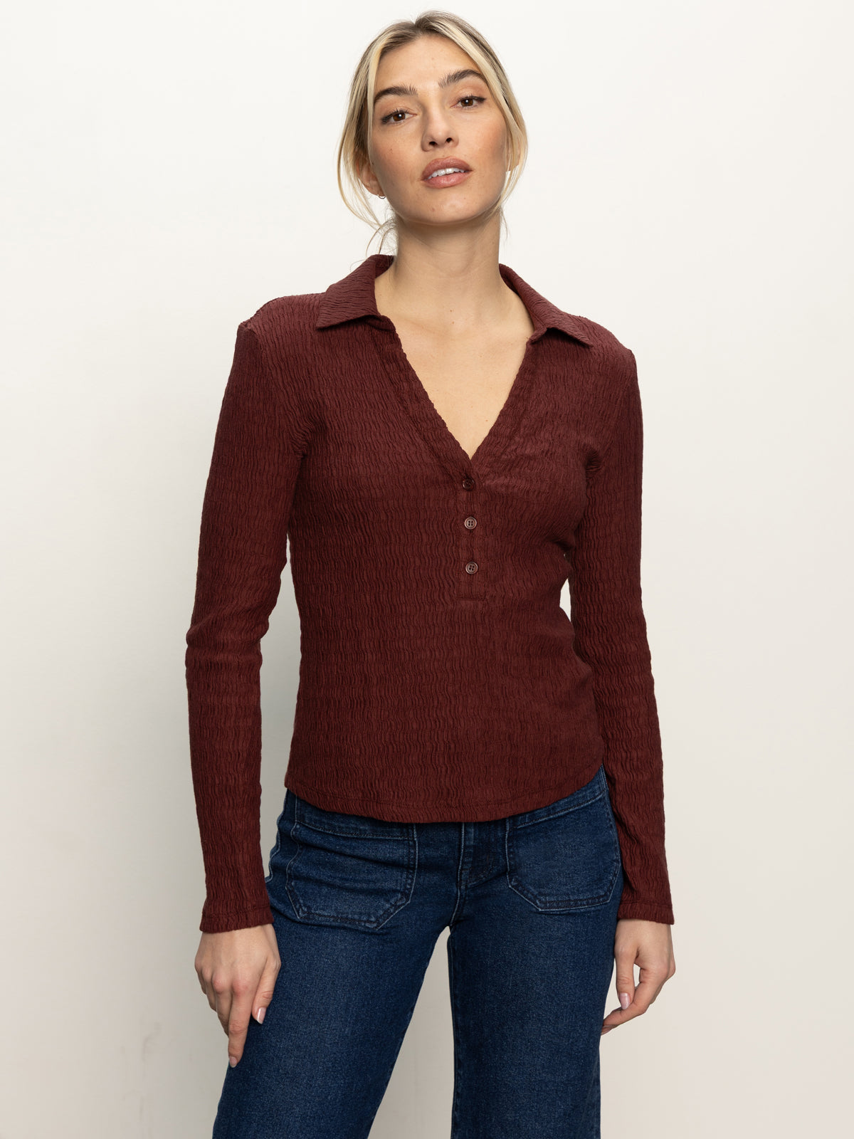 A woman stands against a plain light background, wearing the Sanctuary Clothing long sleeve textured top in chicory and blue jeans. She looks relaxed, with her left hand by her side and her right hand slightly forward.