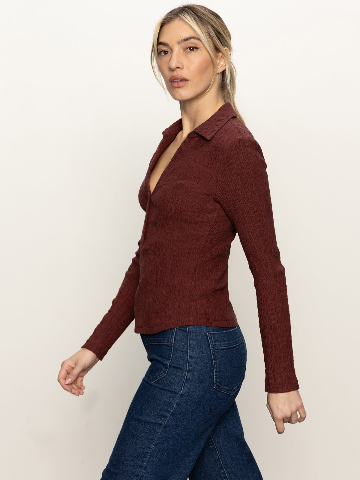 A woman with long blonde hair in a low ponytail wears the Sanctuary Clothing long sleeve textured top chicory with dark blue jeans, walking and looking at the camera against a plain light background.