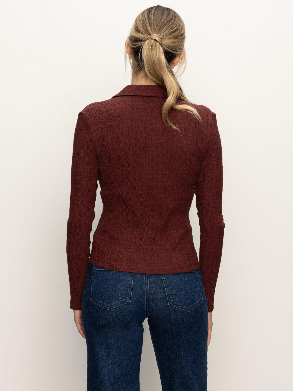 A woman with blond hair in a low ponytail wears the Sanctuary Clothing long sleeve textured top in chicory and blue jeans, facing away from the camera against a plain light background.