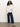 A woman with curly hair wears the Sanctuary Clothing long sleeve textured top in chalk, paired with blue wide-leg jeans and tan flats, standing against a light background with her arms crossed and looking forward.