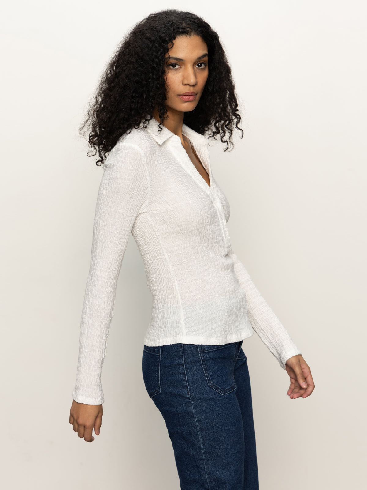 A person with long curly hair wears the Sanctuary Clothing long sleeve textured top in chalk with blue jeans, standing against a plain light background and looking at the camera with a neutral expression.