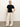 A person stands indoors against a plain background, wearing the Sanctuary Clothing shirred waist dolman tee in black, wide-leg white pants, and beige shoes. They have light hair pulled back and one hand in their pocket, with a relaxed expression.