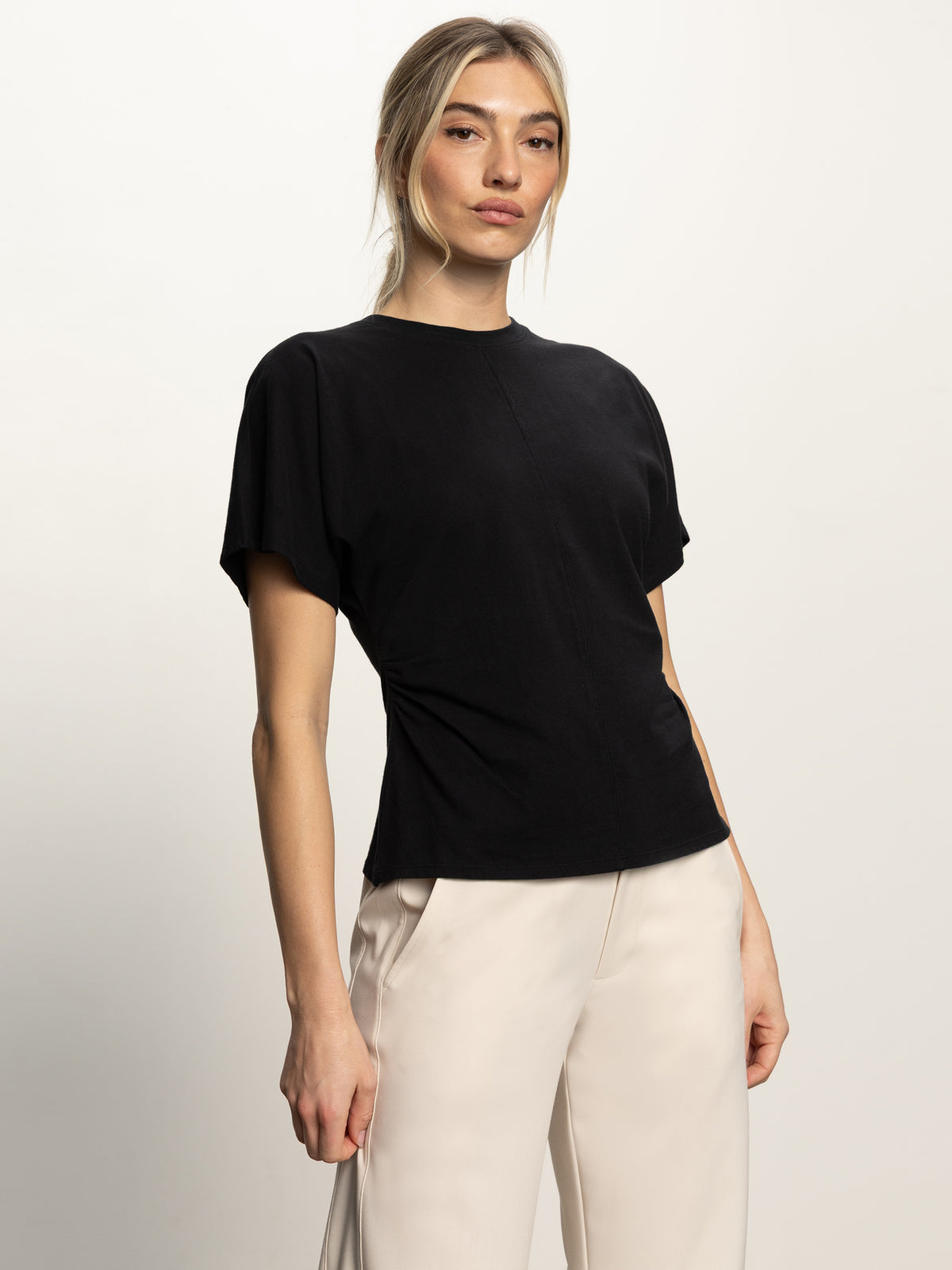 A woman with light hair pulled back wears the Sanctuary Clothing shirred waist dolman tee in black and light-colored pants, standing against a plain light background and looking confidently at the camera.