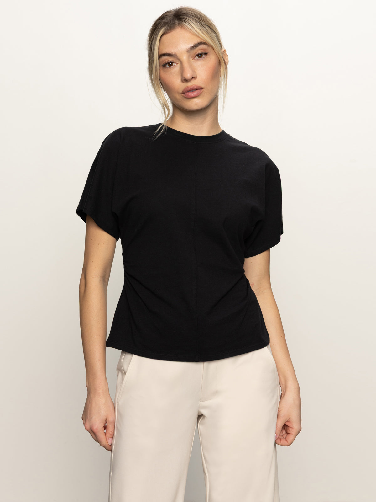 A woman with blonde hair tied back wears the Sanctuary Clothing shirred waist dolman tee in black, paired with light beige pants, standing against a plain light background and looking directly at the camera with a neutral expression.