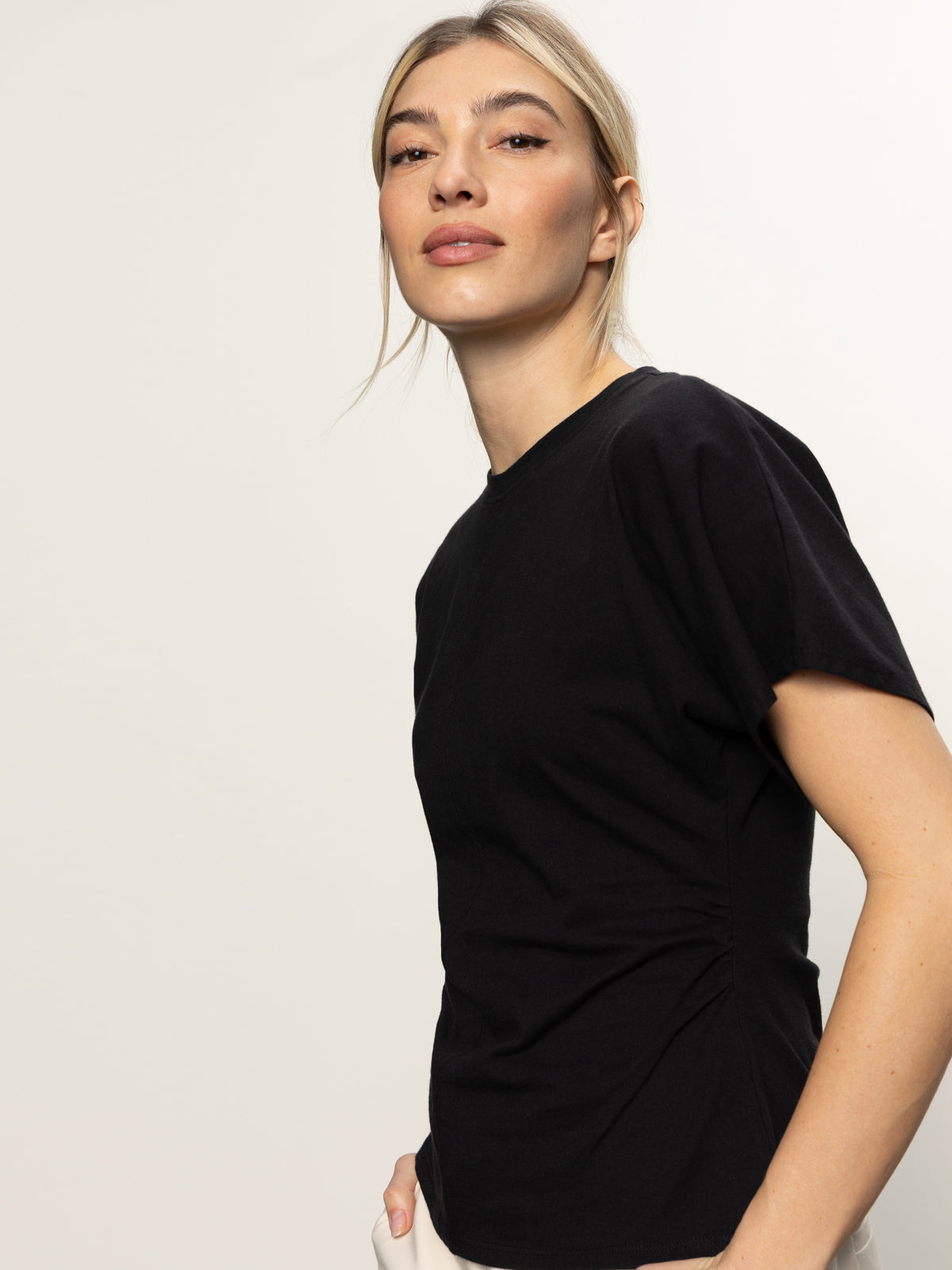 A woman with light skin and blonde hair confidently poses against a plain light background in a Sanctuary Clothing shirred waist dolman tee in black, paired with white pants.