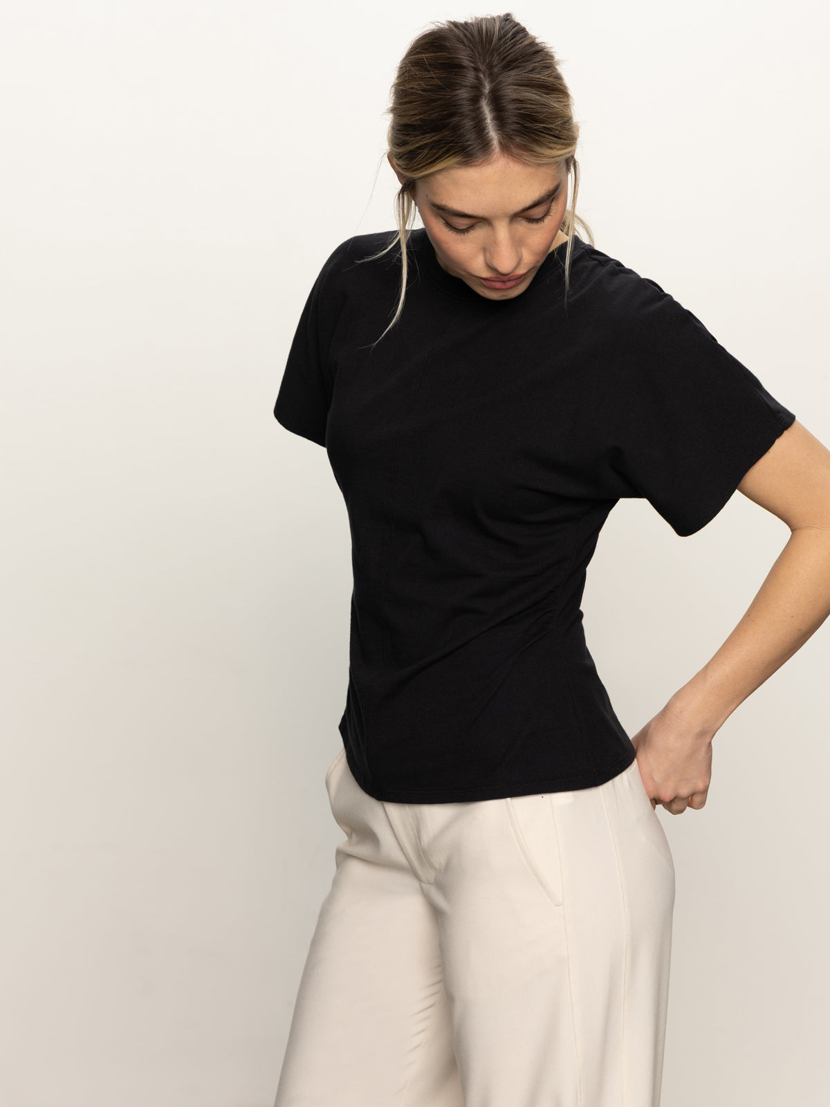 A woman with blonde hair looks down while adjusting her light-colored pants. She is wearing the Sanctuary Clothing shirred waist dolman tee in black, standing against a plain, light background.