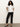 A woman with curly hair wears the Sanctuary Clothing shirred waist dolman tee in white, paired with black straight-leg pants and black low-heeled shoes, standing against a light background with her hands on her hips.