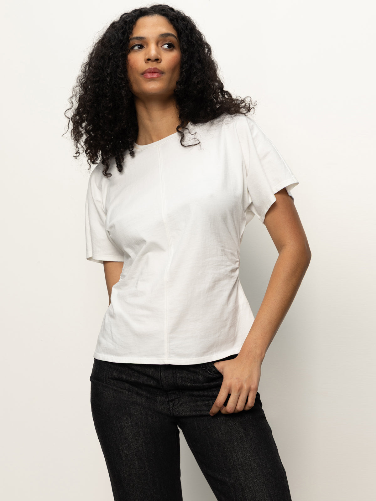 A woman with long curly hair wears the Sanctuary Clothing shirred waist dolman tee in white with dark jeans, standing against a plain light background and looking slightly to the side with one hand in her pocket.