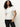 A woman with long curly hair wears the Sanctuary Clothing shirred waist dolman tee in white with dark jeans, standing against a plain light background and looking slightly to the side with one hand in her pocket.