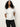 A woman with long curly hair wears the Sanctuary Clothing shirred waist dolman tee in white and dark jeans, standing against a plain light background with her hands behind her back.
