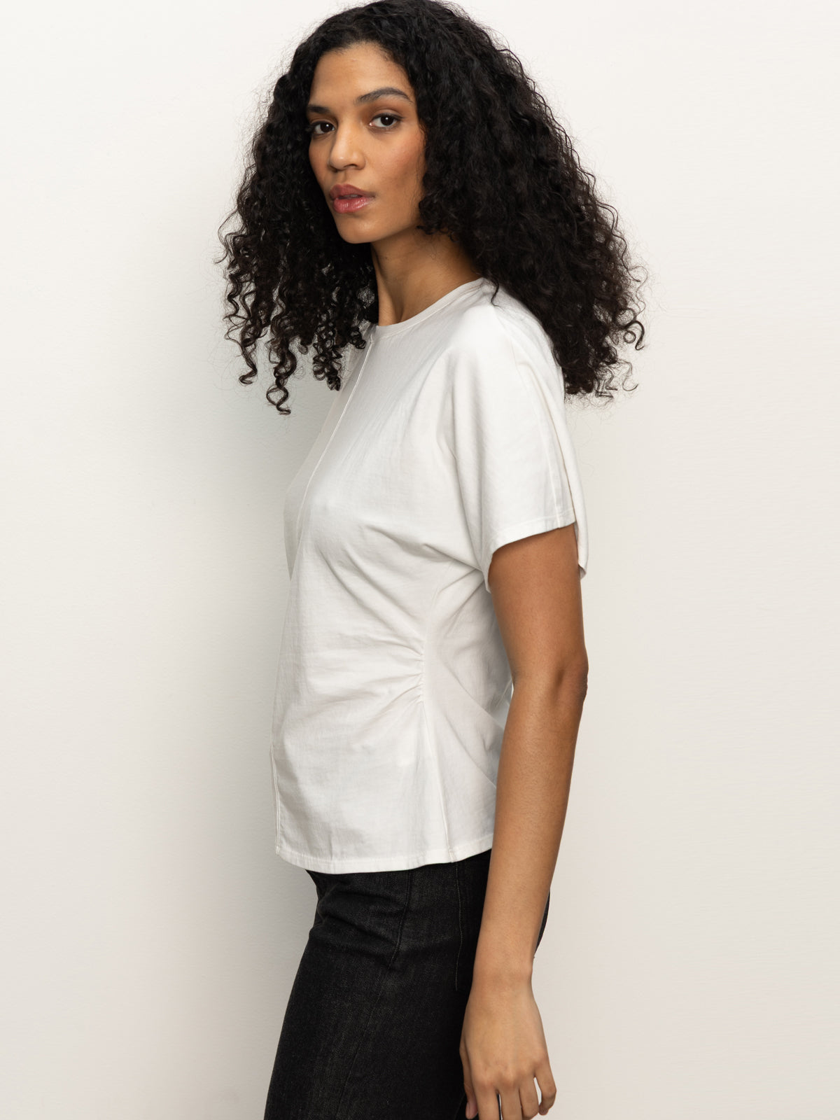 A woman with curly dark hair wears the Shirred Waist Dolman Tee in white by Sanctuary Clothing and dark pants, standing sideways against a plain light background.