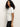 A woman with curly dark hair wears the Shirred Waist Dolman Tee in white by Sanctuary Clothing and dark pants, standing sideways against a plain light background.