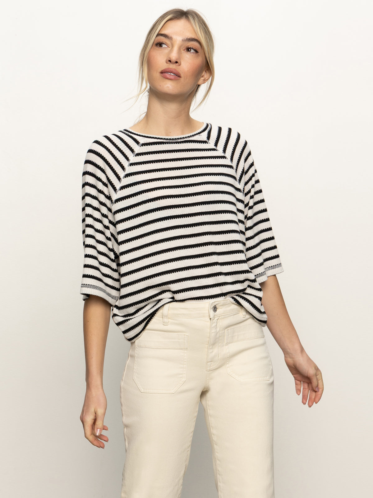 A woman stands against a white background wearing the Sanctuary Clothing boxy raglan tee in black with light oat stripe and high-waisted off-white pants, looking slightly to the side with a relaxed expression.