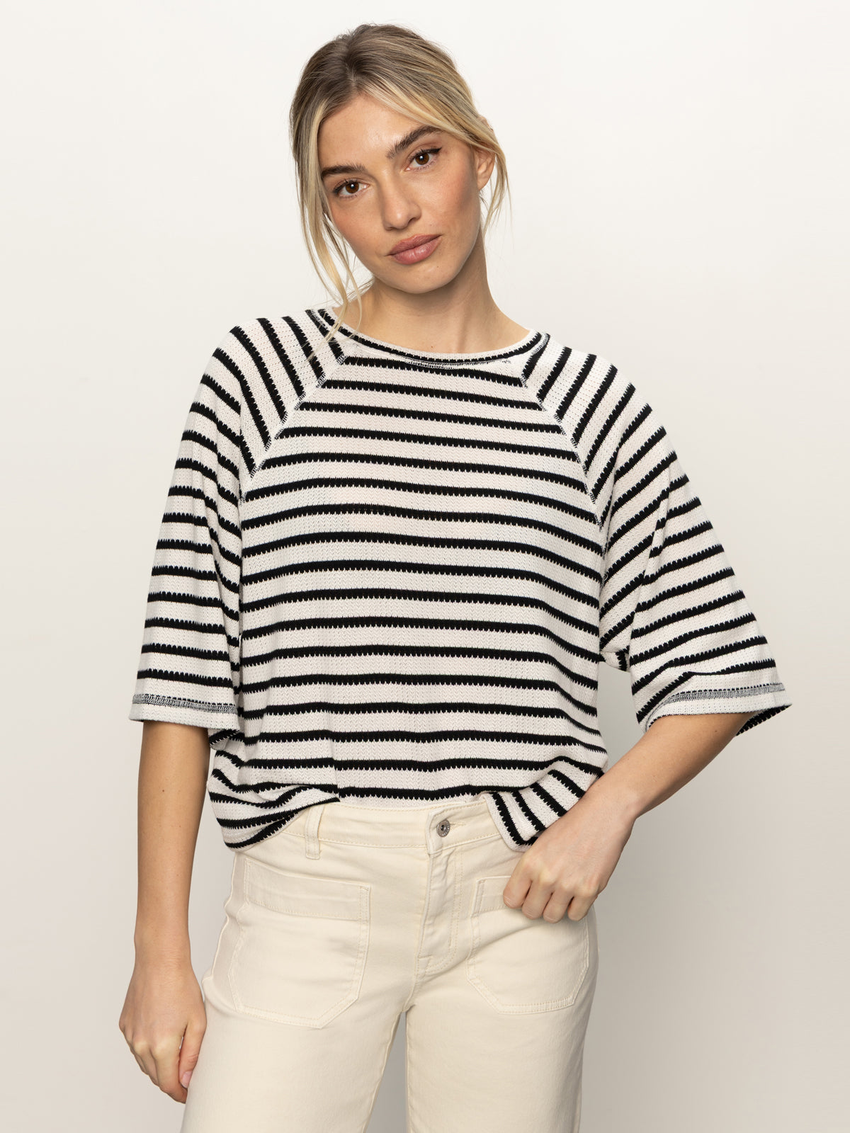 A blonde person stands against a plain light background, facing the camera neutrally, wearing Sanctuary Clothing's boxy raglan tee in black with light oat stripes and light-colored pants.
