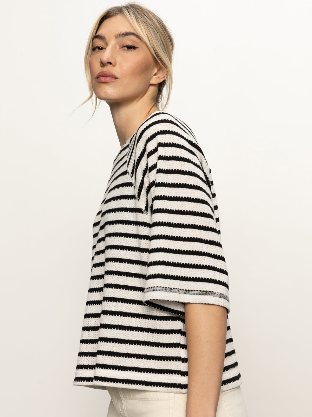 A woman with blonde hair in a loose updo poses against a white background, wearing Sanctuary Clothing’s boxy raglan tee in black with light oat stripe and light-colored pants.