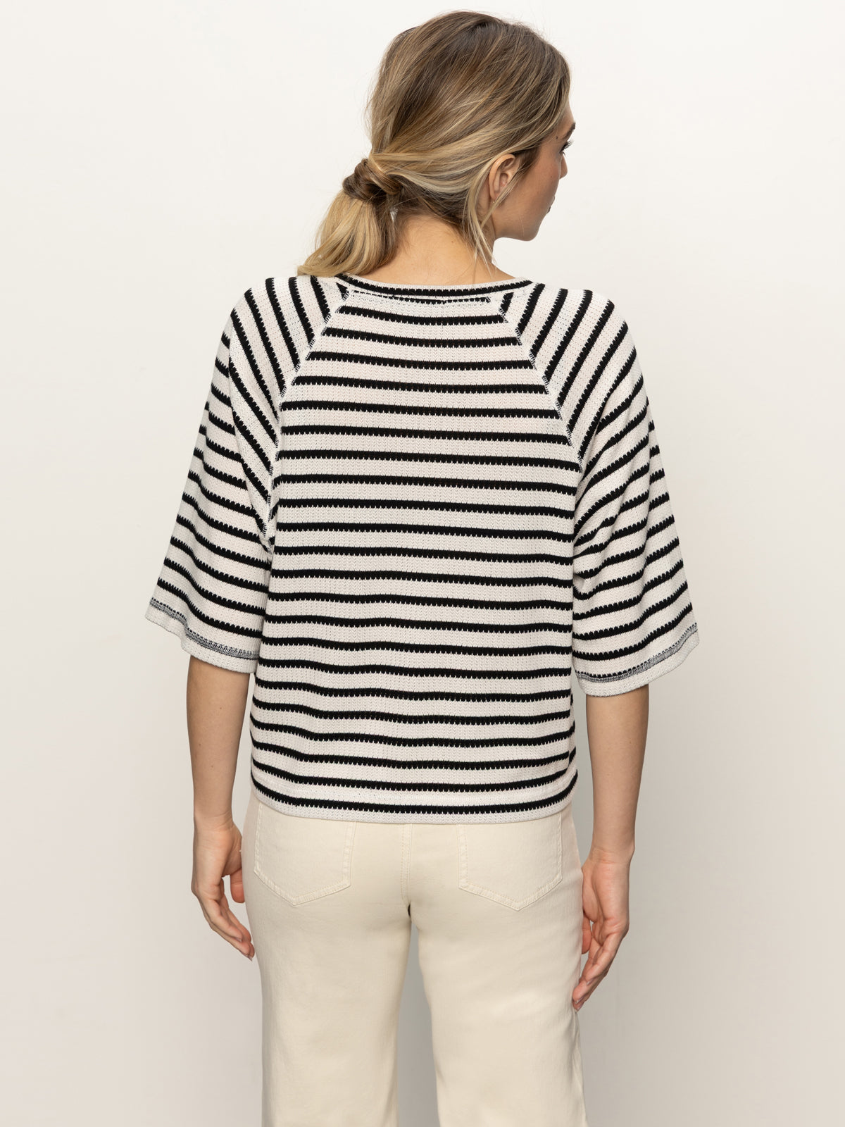 A woman with blonde hair in a low ponytail is wearing the Sanctuary Clothing boxy raglan tee in black with light oat stripe, standing facing away from the camera against a plain background.