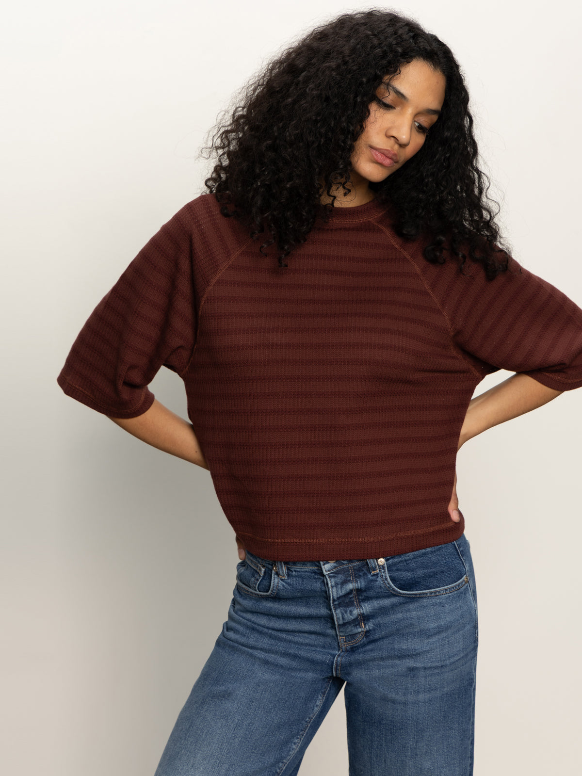 A person with long curly hair wears Sanctuary Clothing's boxy raglan tee in chicory and blue jeans, standing against a plain light background with hands on hips and head slightly tilted down.