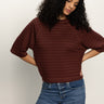 A person with long curly hair wears Sanctuary Clothing's boxy raglan tee in chicory and blue jeans, standing against a plain light background with hands on hips and head slightly tilted down.